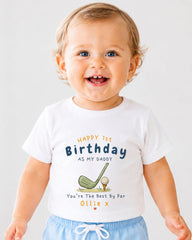A smiling baby boy wearing a white t-shirt with a golf-themed design and text that reads "Happy 1st Birthday as my Daddy You're The Best By Par Ollie x".