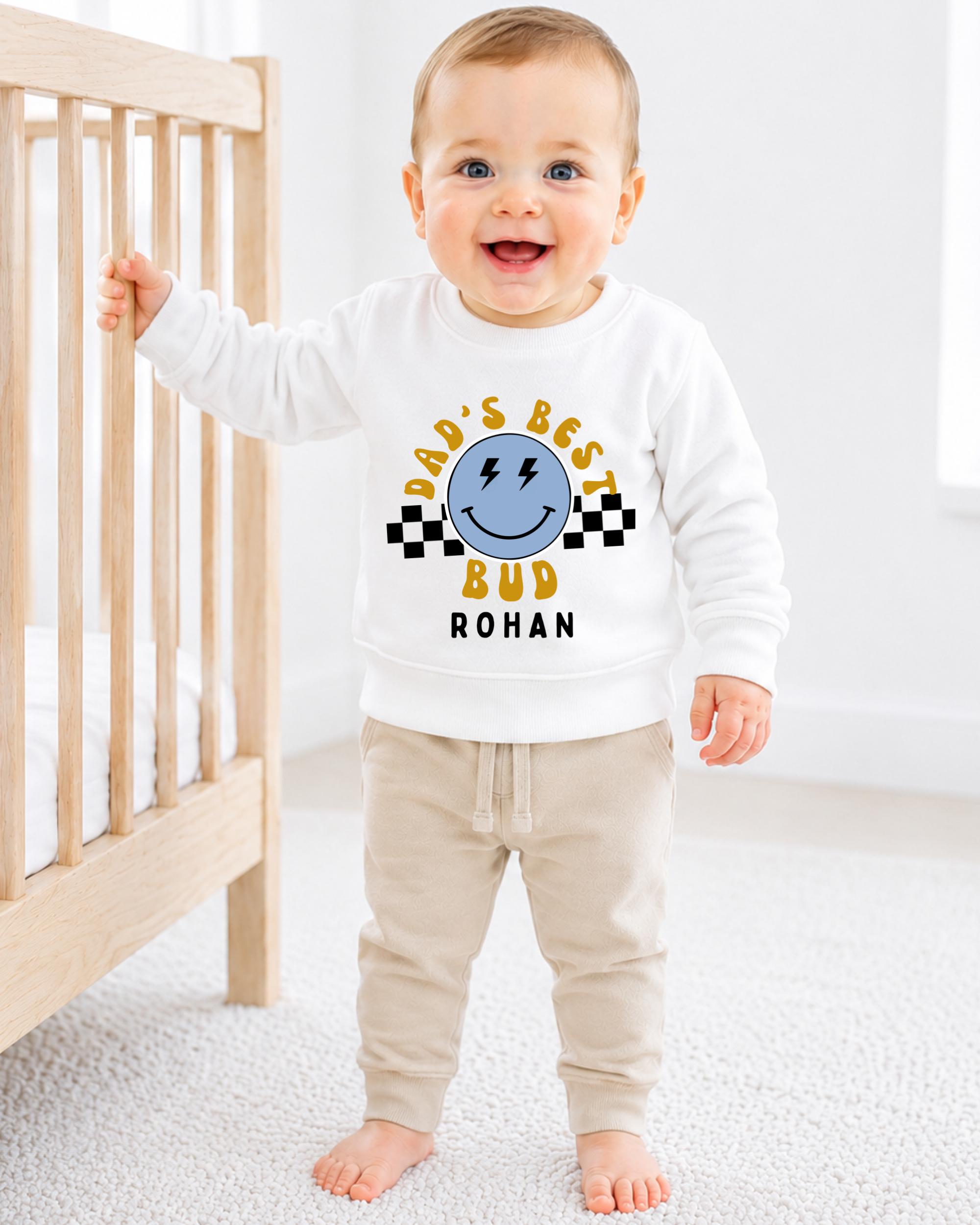 A smiling baby boy wearing a white sweatshirt with a graphic design and text, standing in front of a wooden crib.