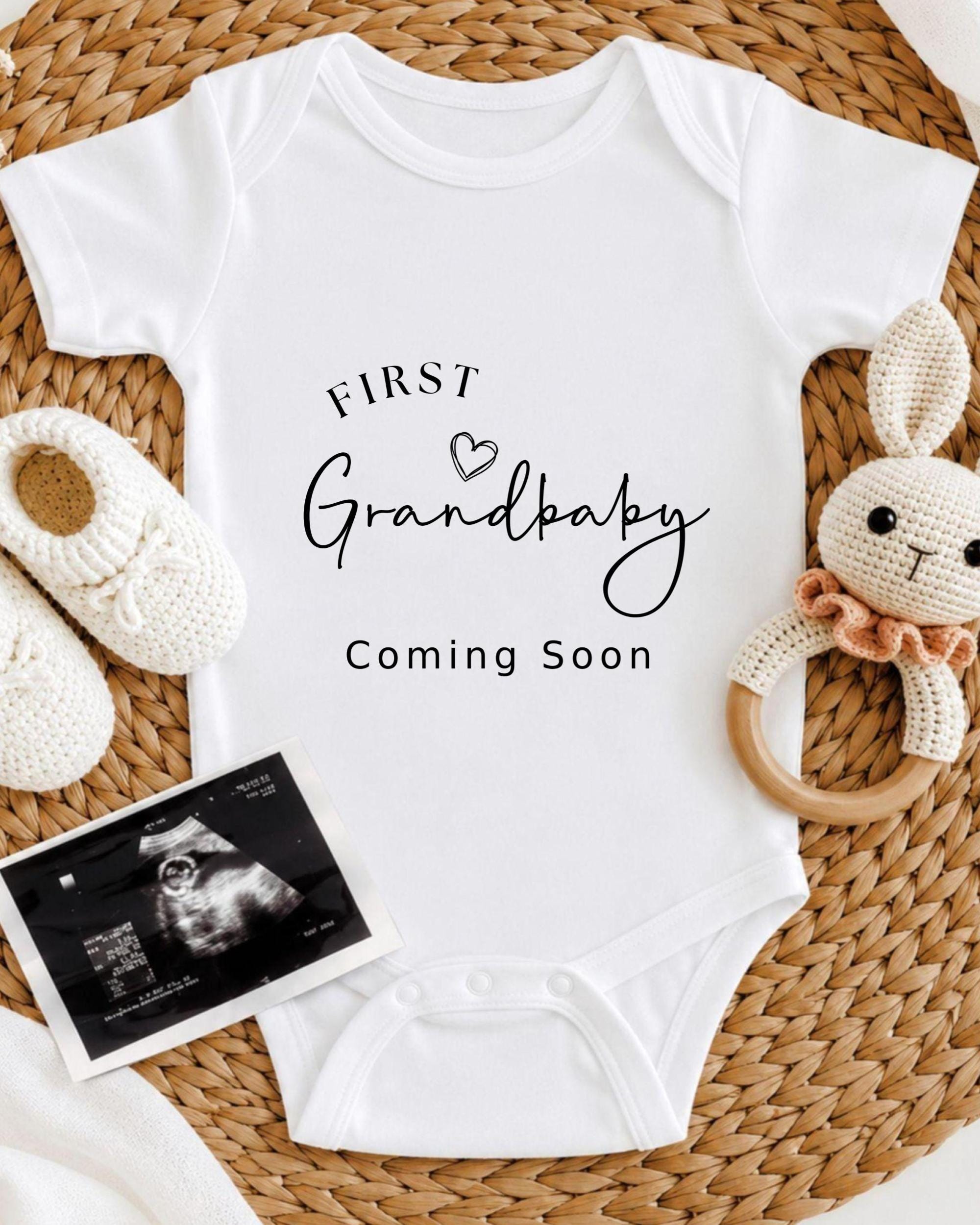 A white onesie with the text "FIRST Grandbaby Coming Soon" printed on it, along with a crocheted rabbit toy and a black and white photograph of an infant's belly.