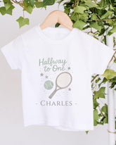 A white t-shirt with a tennis racket and ball graphic printed on it, along with the text "Halfway to One - Charles" printed below.