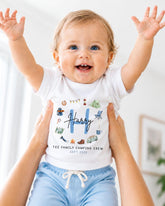 A smiling baby wearing a white shirt with a personalized design and text, being held up by someone's arms.