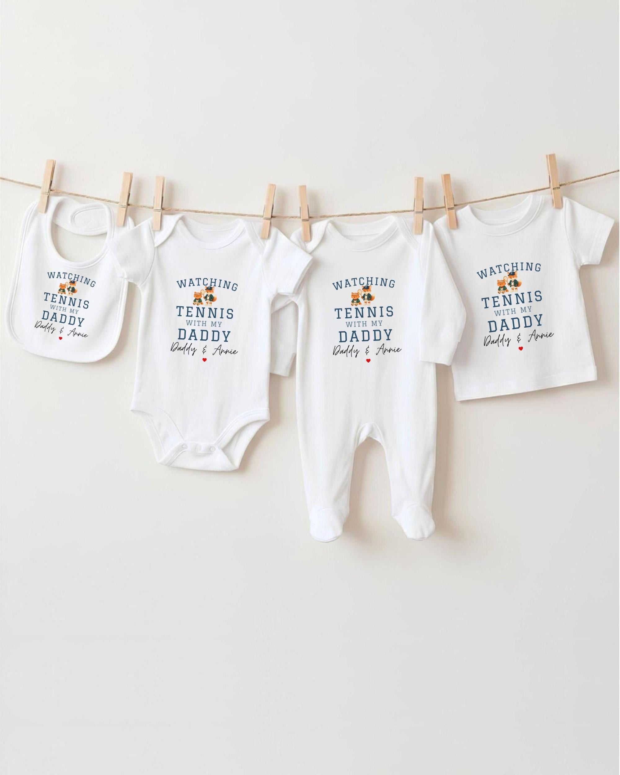 A row of four white baby clothes with the text "WATCHING TENNIS DADDY" printed on them, hanging on a clothesline.