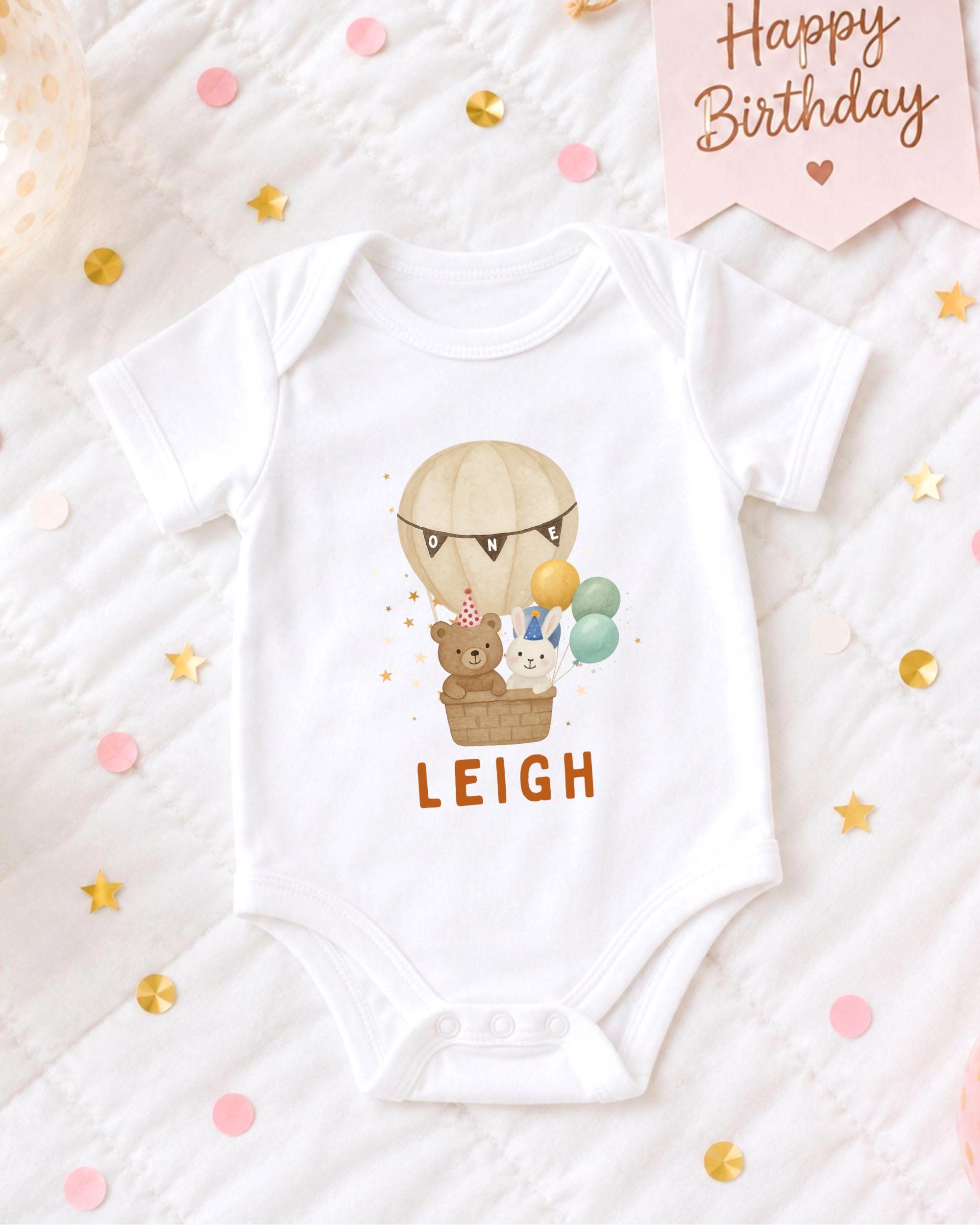 A white baby onesie with a cartoon image of a hot air balloon and animals, along with the name "LEIGH" printed on it, is displayed on a bed with confetti.