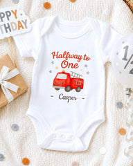 A white onesie with a red fire truck graphic and the text "Halfway to One Casper" printed on it, along with a "Happy Birthday" sign and a gift box.