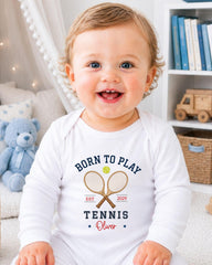 A smiling baby boy wearing a white onesie with a tennis racket and ball design, sitting in a room with a teddy bear and bookshelf in the background.