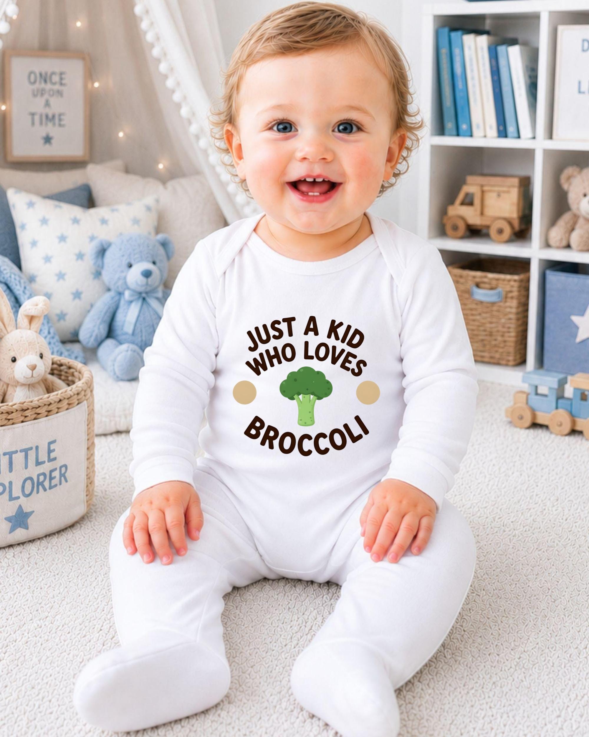A smiling baby boy wearing a white onesie with the text "Just a kid who loves broccoli" is sitting on the floor in a nursery setting.