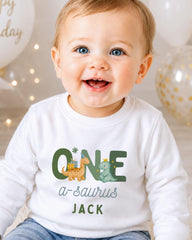 A smiling baby boy wearing a white shirt with a dinosaur and the text "ONE a-saurus JACK" printed on it.
