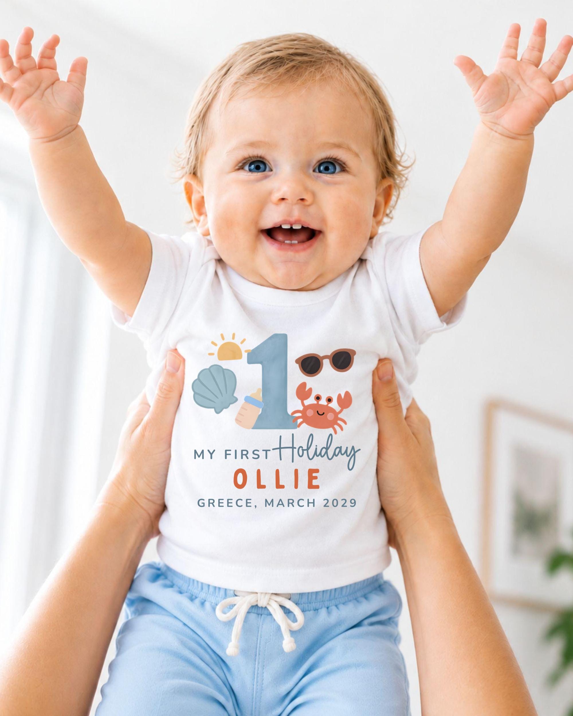 A smiling baby wearing a white t-shirt with a personalized design and text, being held up by someone's arms.