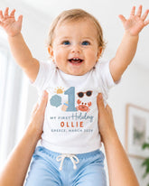 A smiling baby wearing a white t-shirt with a personalized design and text, being held up by someone's arms.