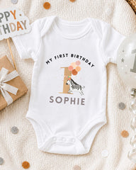 A white onesie with a zebra and the text "My first birthday Sophie" printed on it, along with a "1" and a birthday sign.