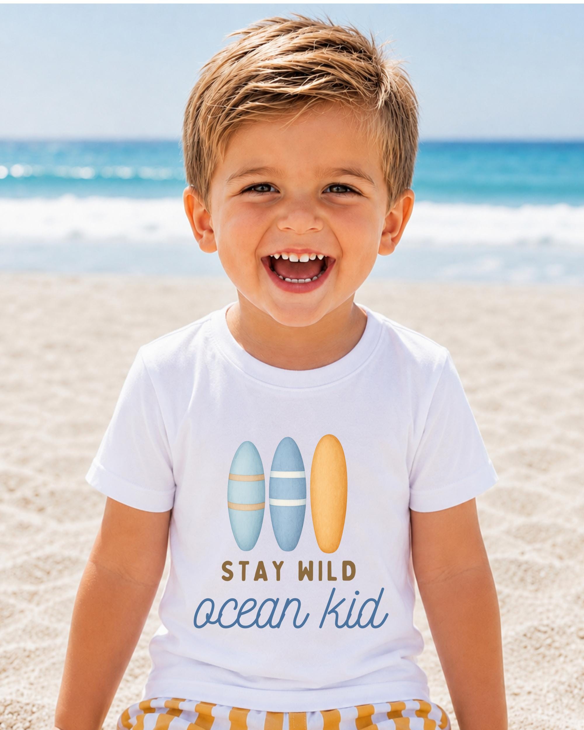 A young boy with a big smile on his face, wearing a white t-shirt with a graphic design of surfboards and the text "STAY MILD ocean kid" printed on it, standing on a sandy beach with the ocean in the background.