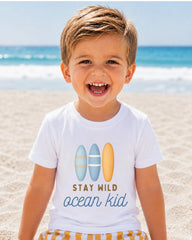 A young boy with a big smile on his face, wearing a white t-shirt with a graphic design of surfboards and the text "STAY MILD ocean kid" printed on it, standing on a sandy beach with the ocean in the background.