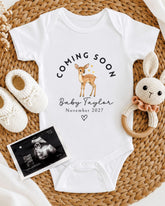 A white onesie with a deer graphic and the text "Coming soon Baby Taylor November 2027" is displayed on a woven placemat, accompanied by a crocheted rabbit toy and a baby shoe.