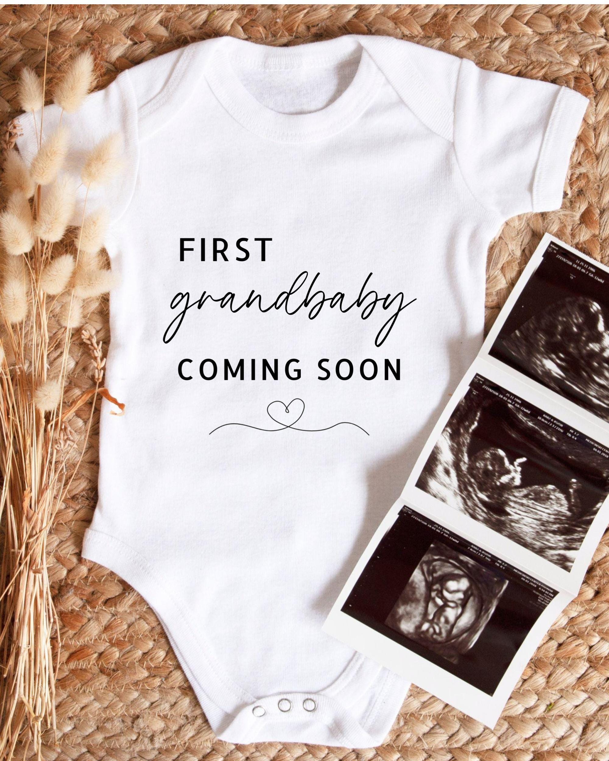 A white baby onesie with the text "FIRST grandbaby COMING SOON" printed on it, along with a heart symbol. The onesie is placed next to three black and white images of an ultrasound of a baby, suggesting the image is a collage of baby images.