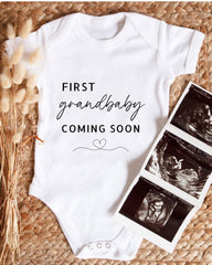A white baby onesie with the text "FIRST grandbaby COMING SOON" printed on it, along with a heart symbol. The onesie is placed next to three black and white images of an ultrasound of a baby, suggesting the image is a collage of baby images.