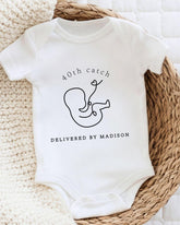 A white baby onesie with a graphic of a baby and text that reads "40th catch DELIVERED BY MADISON".