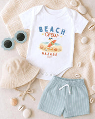 A white onesie with a beach crew design, a beige bucket hat, and a pair of light blue shorts, along with sunglasses and seashells, are arranged on a beige surface.
