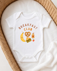 A white onesie with the text "BREAKFAST CLUB" printed on it, along with various breakfast-related icons such as a banana, pretzel, apple, and a jar of jam.