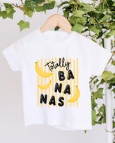 A white t-shirt with a playful graphic design featuring the text "Totally BANANA" and three bananas.