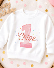 A white sweatshirt with a pink number one and the name "Chloe" printed on it, surrounded by confetti.