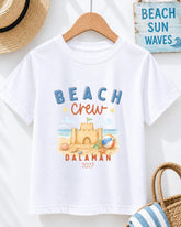 A white t-shirt with a beach-themed design, including a castle, beach ball, and sandcastle, along with the text "BEACH CREW DALAMAN 2027".