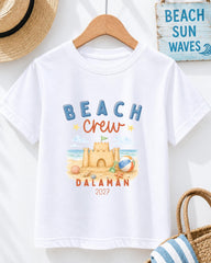 A white t-shirt with a beach-themed design, including a castle, beach ball, and sandcastle, along with the text "BEACH CREW DALAMAN 2027".