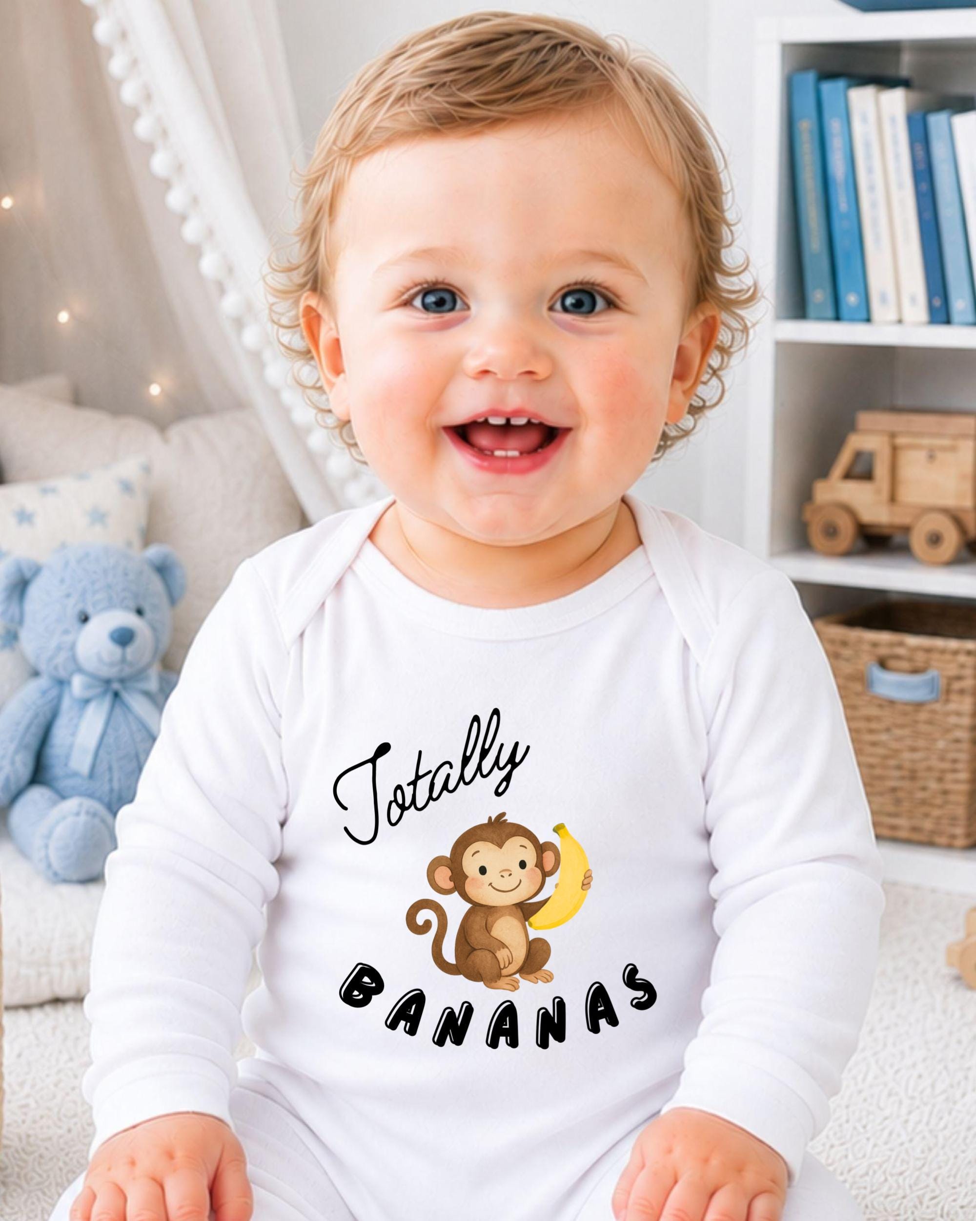 A smiling baby wearing a white onesie with a monkey and bananas printed on it, sitting in a room with a teddy bear and bookshelf in the background.