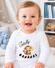 A smiling baby wearing a white onesie with a monkey and bananas printed on it, sitting in a room with a teddy bear and bookshelf in the background.
