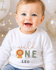 A smiling baby boy wearing a white shirt with a lion graphic and the text "wild one leo" printed on it.