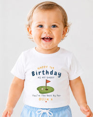 A smiling baby boy wearing a white t-shirt with a golf hole graphic and text that reads "Happy 1st Birthday as my Daddy You're The Best By Par Ollie x".