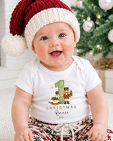 A smiling baby wearing a red and white Santa hat and a white t-shirt with a Christmas-themed design.