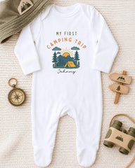 A white onesie with a camping-themed design, a compass, a hat, and a pair of binoculars, all arranged on a beige surface.