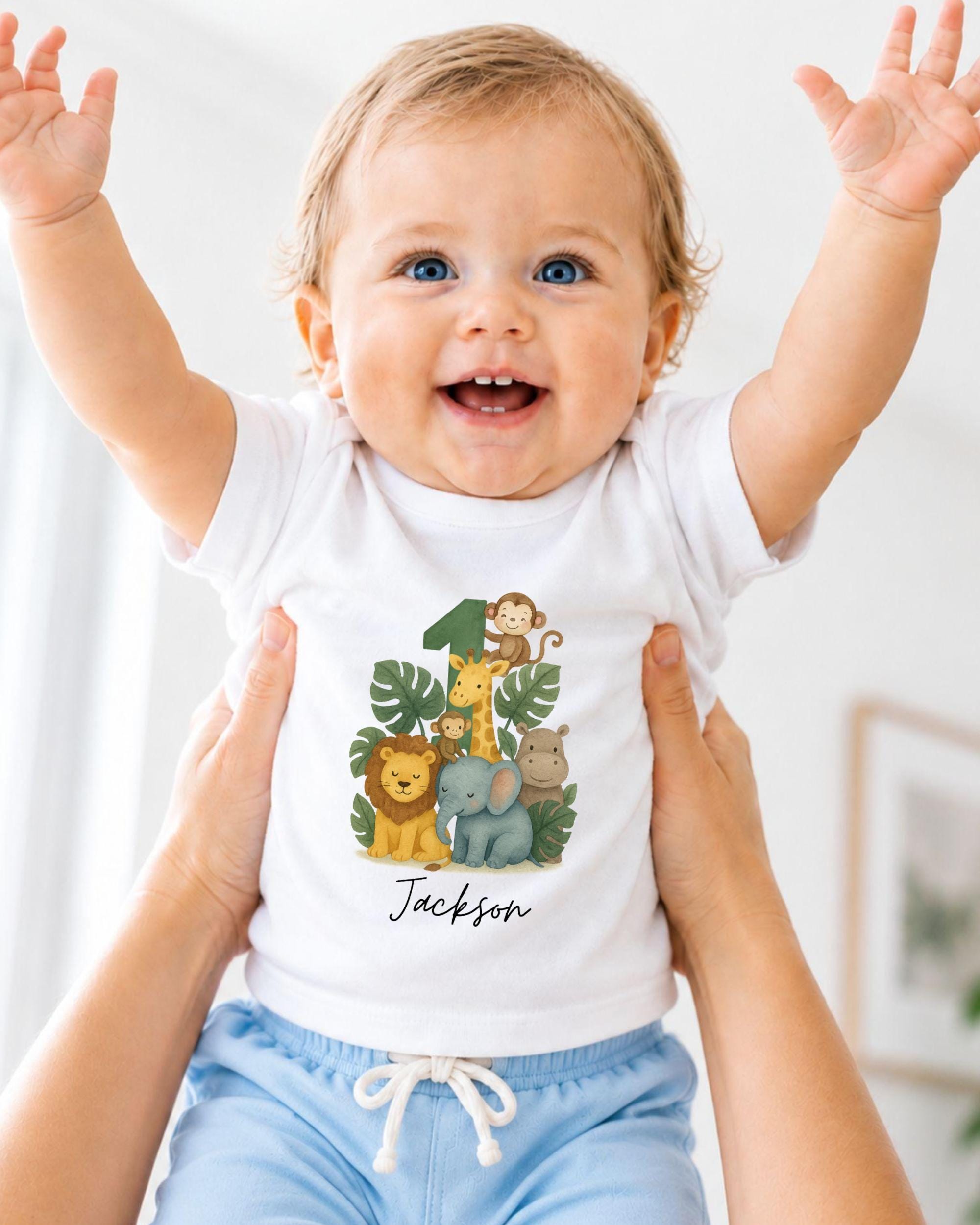 A smiling baby wearing a white t-shirt with a jungle-themed graphic and the name "Jackson" printed on it, being held up by someone's arms.