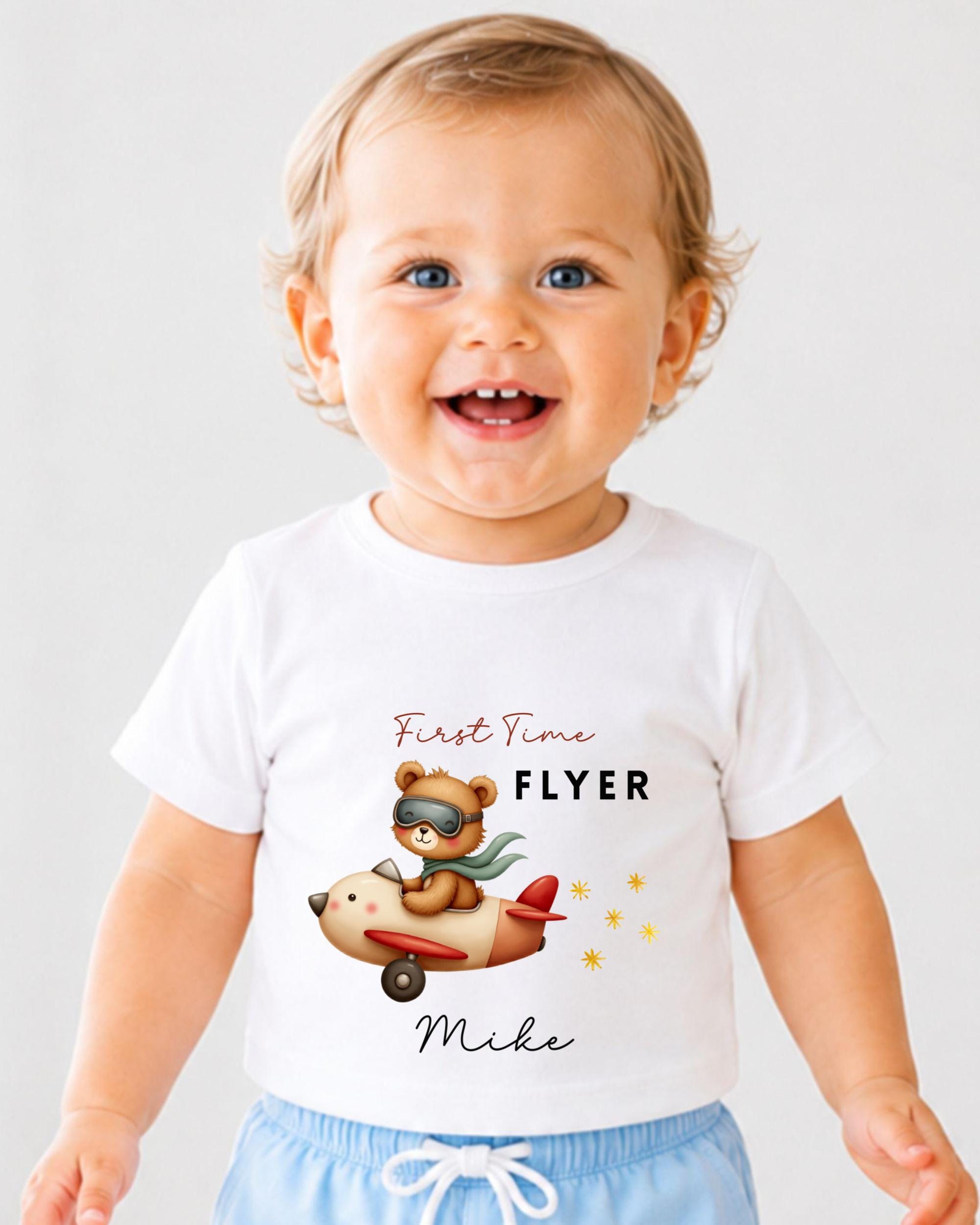 A smiling baby wearing a white t-shirt with a cartoon image of a bear piloting a plane and the text "First Time Flyer Mike" printed on it.