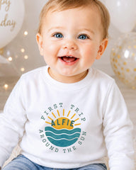 A smiling baby wearing a white shirt with a graphic design and text.