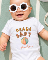 A smiling baby wearing a white onesie with the text "BEACH BABY" and a beach ball graphic, along with sunglasses.