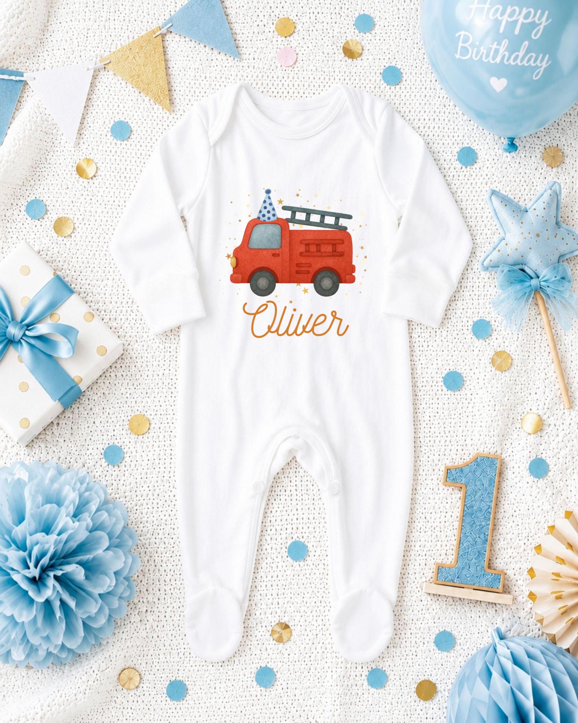 A white onesie with a red firetruck design, surrounded by birthday decorations and confetti.