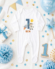 A white onesie with the number 1 and a space suit printed on it, surrounded by birthday decorations such as a blue balloon, blue and gold confetti, and a blue and white polka dot gift box.