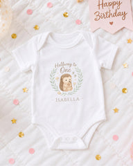 A white onesie with a hedgehog design and the text "Halfway to One" printed on it, along with the name "Isabella" below the hedgehog image. The onesie is placed on a bed with confetti and a "Happy Birthday" sign.