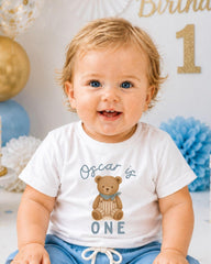 A smiling baby boy wearing a white t-shirt with a teddy bear and the text "Oscar is 1" is sitting in front of a birthday party backdrop.