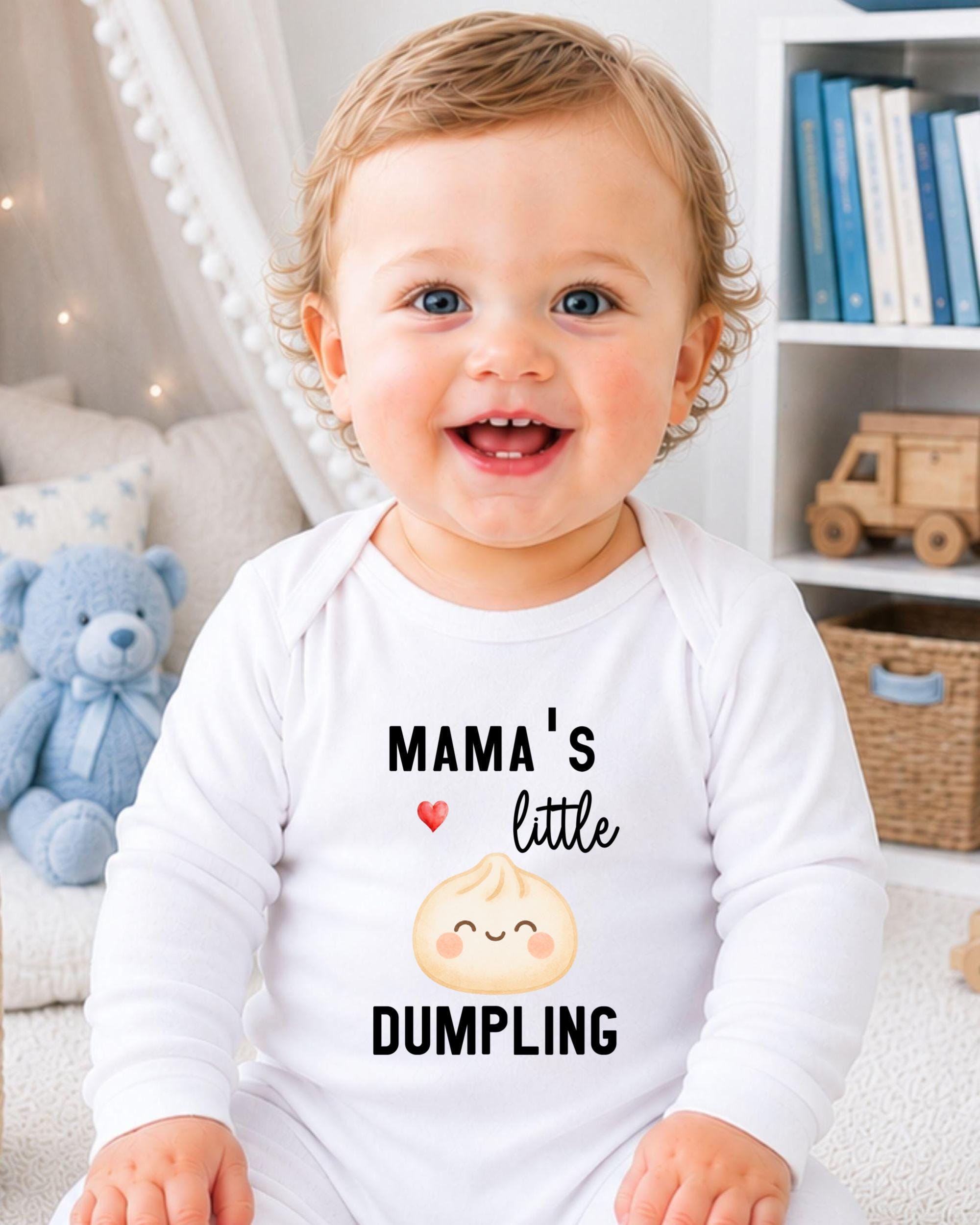 A smiling baby wearing a white onesie with a design of a smiling dumpling and the text "Mama's little DUMPLING" printed on it.
