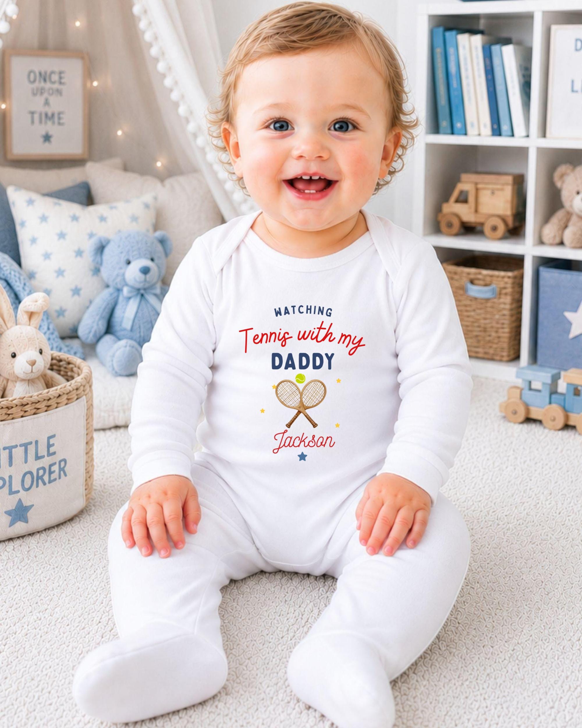 A smiling baby boy wearing a white onesie with tennis and his name printed on it, sitting on a carpeted floor in a room with a bookshelf and stuffed animals in the background.