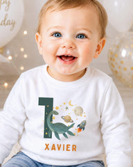 A smiling baby boy wearing a white shirt with a dinosaur and space-themed design, sitting in front of balloons.