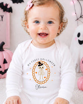 A smiling baby girl wearing a white onesie with a Halloween-themed design and text.
