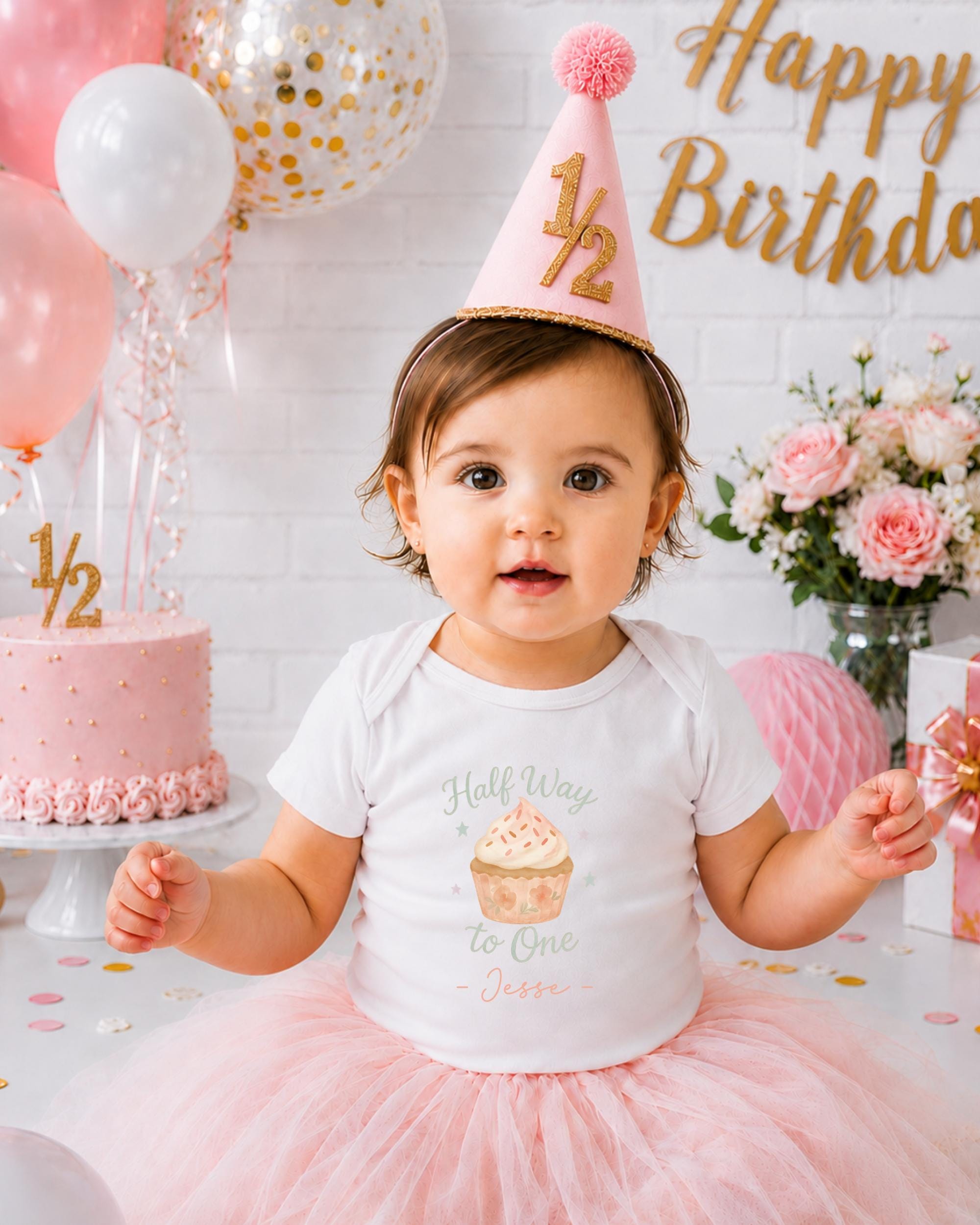 A young girl wearing a birthday hat and a white t-shirt with a cupcake design is sitting in front of a birthday cake and balloons.