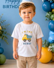 A young boy wearing a white t-shirt with a truck graphic and the text "I'm two Henry" stands in front of a blue background with balloons.