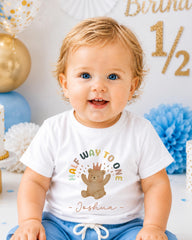 A smiling baby boy wearing a white t-shirt with a cartoon bear and the text "Halfway to one" is sitting in front of a birthday party backdrop.