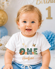 A smiling baby boy wearing a white t-shirt with a dinosaur and the name "Hudson" printed on it, sitting in front of a birthday party backdrop.