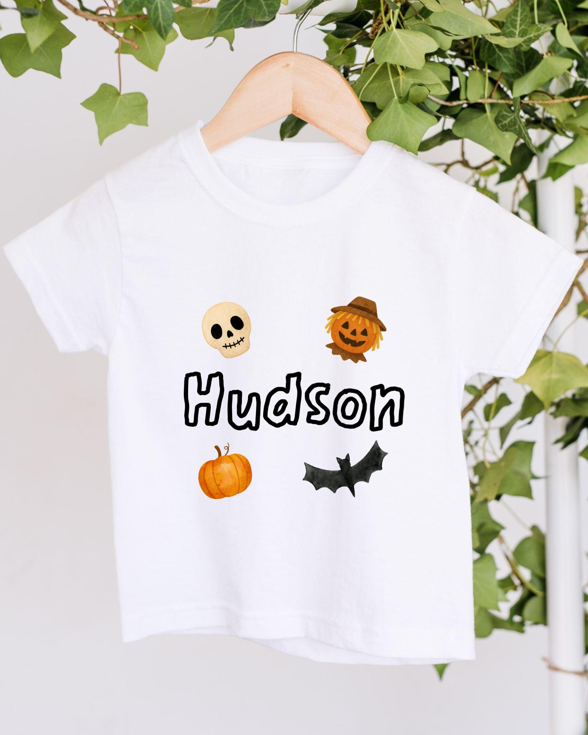 A white t-shirt with a Halloween-themed design, including a skull, a scarecrow, a pumpkin, and a bat.