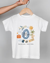 A white t-shirt with a graphic design featuring various travel-related icons, including a suitcase, a plane, a palm tree, and a sun. The text on the t-shirt says "Barcelona, July 2023".
